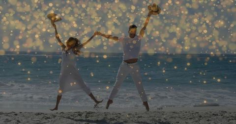 Sunlit couple jumping on ocean shore holding straw hats, celebrating joy with golden bokeh