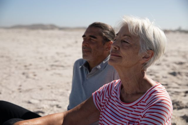 Senior Caucasian couple enjoying time at the beach on a sunny day, sitting on the beach, with sand and blue sky in the background. Summer tropical beach vacation.