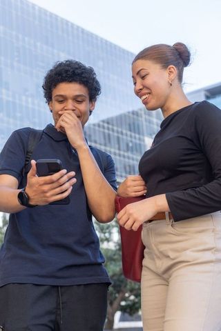 Smiling african american friends checking smartphone outside mirrored glass office
