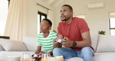 African american father and son playing video games on sofa sharing snacks at home