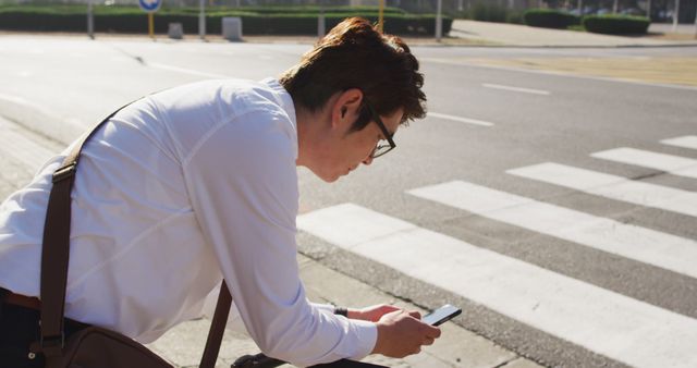 Businessman bending over while checking phone at pedestrian crosswalk on sunny day. Capturing modern office workers' reliance on technology in urban environments. Useful for illustrating business, technology, or transportation themes.