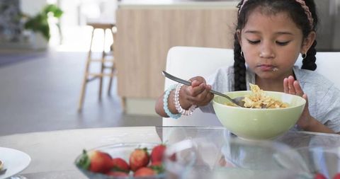 Young girl scooping cereal from pale green bowl at sunlit kitchen table with strawberries