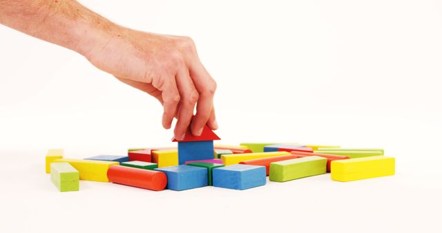 A Caucasian hand is placing a red roof on a small structure made of colorful wooden blocks, with copy space. Building with blocks often symbolizes creativity, planning, and the concept of construction or development in various contexts.
