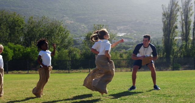 Children of diverse ethnicities are participating in a sack race at a school sports event, with a teacher or coach encouraging them. Sack races are a traditional, fun activity that promote physical fitness and teamwork among young students.