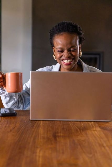 African american woman smiling while working on laptop holding mug at wooden table