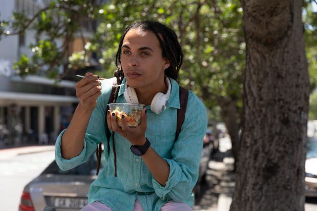 Biracial alternative man with dreadlocks and headphones out and about in the city on a sunny day, eating takeaway lunch. Urban trendy man on the go.