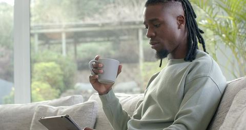 African American man relaxing on sofa holding mug and scrolling tablet by bright window