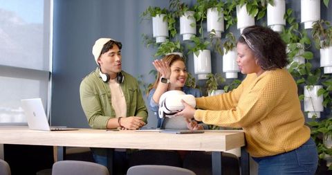 Diverse young professionals sharing vr headset at coworking bar with greenery and laptop