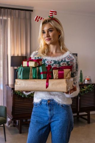 Young woman holding wrapped christmas gifts wearing candy cane headband in cozy living room