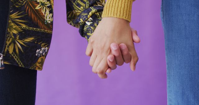 Closeup of two people holding hands against a purple background. This image can be used to depict themes of connection, intimacy, and support, as well as highlighting fashion and stylish clothing. Suitable for relationship advice articles, mental health and wellness promotions, or fashion blogs.