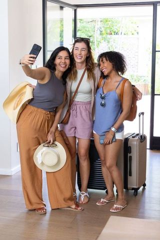 Smiling diverse women taking selfie in sunlit entryway with luggage, ready for vacation