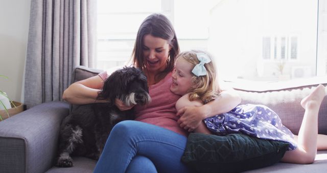 Mother and young daughter bonding while relaxing on the couch with their pet dog. The scene captures a cheerful family moment full of love and companionship. Perfect for content related to family lifestyle, home living, pet care, and happy family moments.