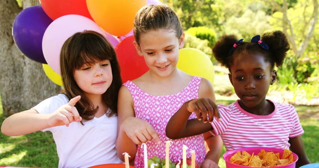 A diverse group of young girls celebrates a birthday outdoors, surrounded by colorful balloons and a cake, with copy space. Their joyful expressions and the festive setting evoke the happiness of childhood celebrations.
