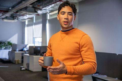 Asian professional standing in open office holding mug and gesturing during discussion