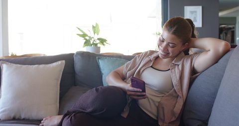 Woman relaxing on sofa in casual loungewear smiling while using smartphone in living room