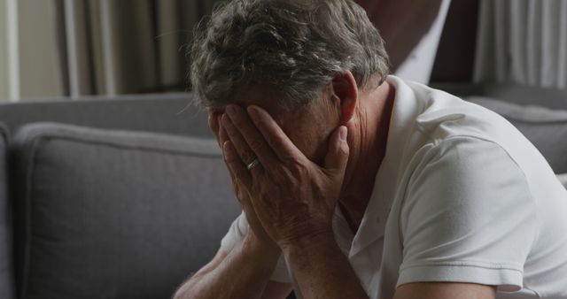 Senior Caucasian man appears distressed at home. His hands cover his face, suggesting worry or sadness in a domestic setting.