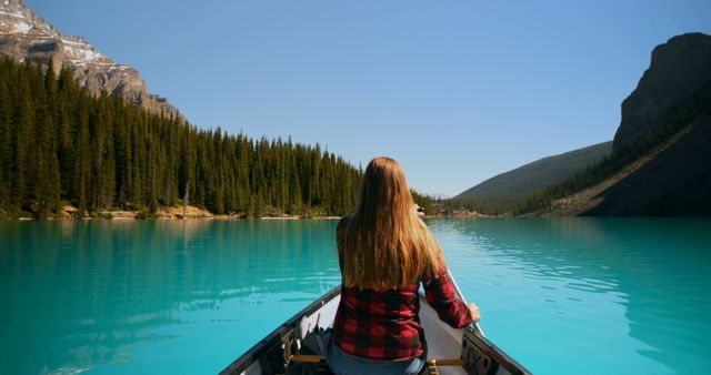 Woman canoeing on a serene turquoise lake surrounded by lush evergreen forest and rugged mountains. Ideal for outdoor adventure, solo travel, and nature exploration themes. Great for content focusing on tranquility, nature activities, summer vacations, and scenic landscapes.