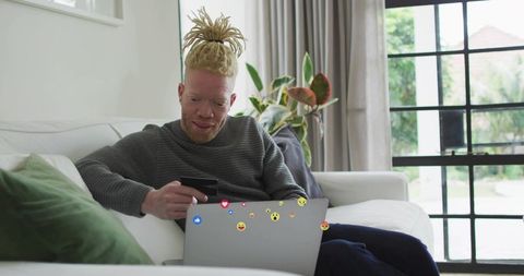 Man relaxing on sofa using smartphone and laptop with social media reactions in bright living room