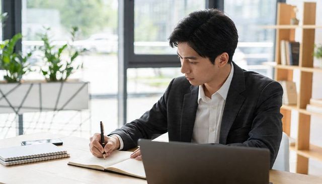 Focused asian businessman writing notes at coworking desk with laptop and notebook