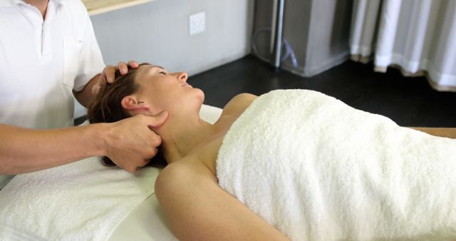 A woman is lying on a massage table, wrapped in a towel, receiving a neck massage from a therapist. This image can be used for content related to spa services, wellness treatments, stress relief, body care, and therapy.
