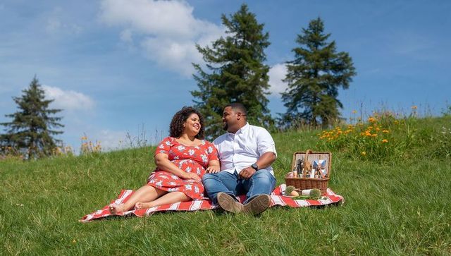 Black couple enjoying picnic on sunlit meadow hillside with wildflowers and wicker basket