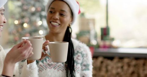 African american friends toasting with cozy ribbed mugs in santa hats by christmas tree