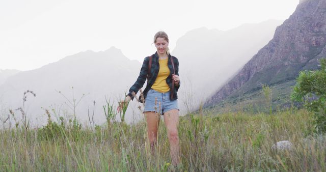 Woman hiking in grassy area with mountains in the background during early morning light. Casual outfit suggests leisure and relaxation, perfect for inspiration on outdoor adventures, travel destinations, and promoting a healthy and active lifestyle.
