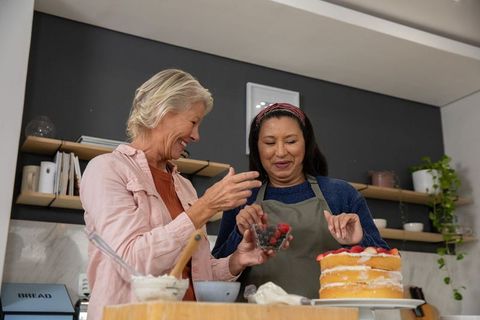 Two women decorating layered cake with fresh berries in cozy kitchen sharing baking joy