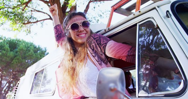 The image shows a young woman leaning out of a classic van window with her arm raised and a joyful expression. She is wearing sunglasses and boho-style clothing, depicting a carefree and adventurous vibe. This image can be used for travel blogs, lifestyle magazines, advertisements promoting road trips, holiday marketing materials, and websites related to adventure and exploration.