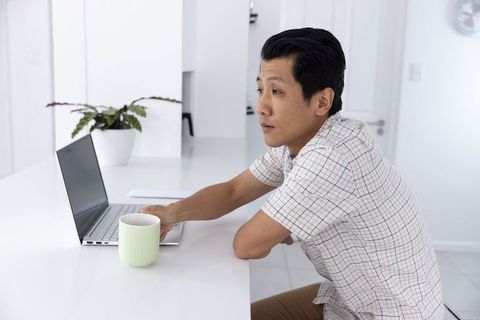 Asian man working at bright minimalist countertop with laptop and mug, home workspace