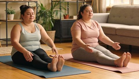 Plus size women practicing mindful breathing on yoga mats in sunlit living room