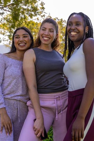 Smiling diverse women posing in sunlit park in athleisure track pants holding smartphone