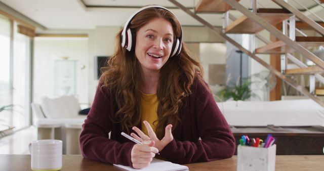 Woman with red hair wearing headphones and a brown cardigan, smiling while enjoying music at home. She is sitting at a wooden table with a coffee mug, stationery, and a notepad, creating a cozy and organized home environment. Ideal for use in content related to home lifestyle, relaxation, music enjoyment, remote work, and everyday life scenes at home.
