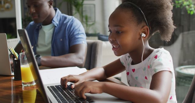 Young girl wearing earphones engaging in online learning on laptop while her father is working on his own laptop. Scene depicts the modern educational environment and work-from-home dynamic. Can be used to illustrate remote education, parent-child bonding, or remote work scenarios.