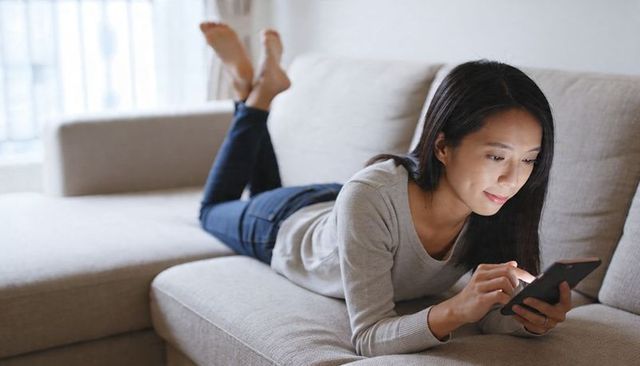 Asian woman relaxing on beige sectional sofa scrolling smartphone in minimalist daylight