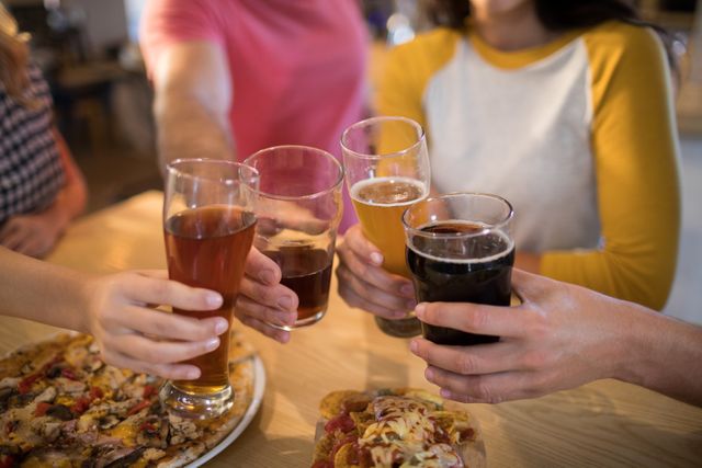 Friends are toasting with various beers at a restaurant table, indicating a casual and fun gathering. The table is filled with food, including pizza, suggesting a relaxed and enjoyable meal. This image can be used for promoting social events, restaurant advertisements, or articles about friendship and socializing.
