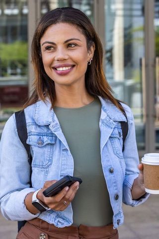 Smiling african descent woman holding coffee and phone outside urban office entrance ready
