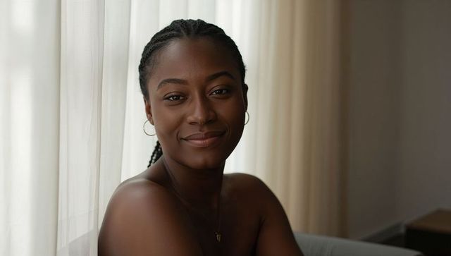 Smiling woman with braids near window wearing gold hoops and pendant in soft natural light