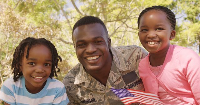 A smiling African American military serviceman is pictured with two happy children, a boy and a girl, outdoors on a sunny day, with copy space. Their joyful expressions and close proximity suggest a strong family bond and the pride they share in their military connection.