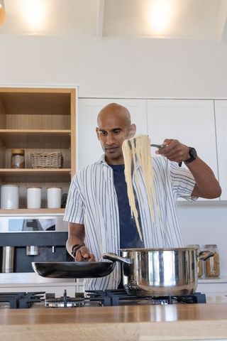 Indian man cooking spaghetti on stovetop in modern kitchen lifting noodles with tongs