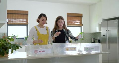 Young women sorting recycling at bright kitchen island labeled glass plastic paper bins