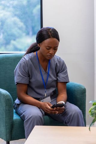 African nurse sitting in scrubs holding smartphone and lanyard, looking worried in staff lounge
