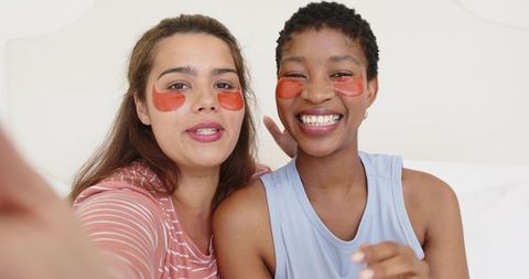 Smiling female friends wearing orange under-eye patches taking selfie on bed