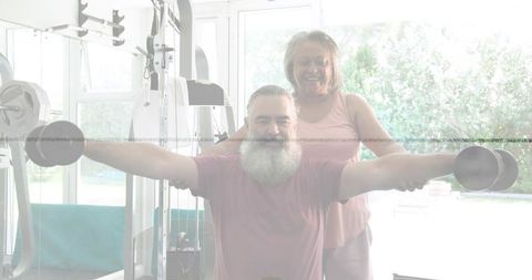 Senior couple doing lateral raises in home gym, woman spotting partner during workout