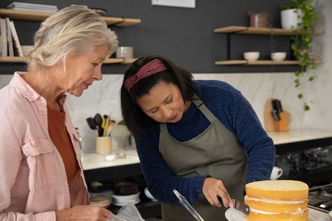 Senior woman teaching cake decorating to adult woman while icing layered cake in home kitchen