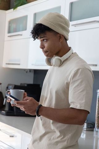 African american man standing in kitchen holding mug and smartphone wearing headphones and beanie