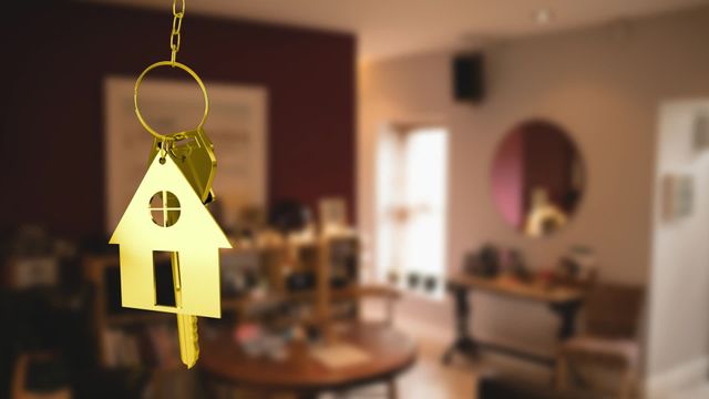 Golden key with house symbol hangs in focus, background shows cozy home interior with furniture blurred. Indicates concept of moving to a new home or settling in. Ideal for real estate promotions, blogs on moving tips, home decor inspiration, advertisements related to new beginnings and home ownership.