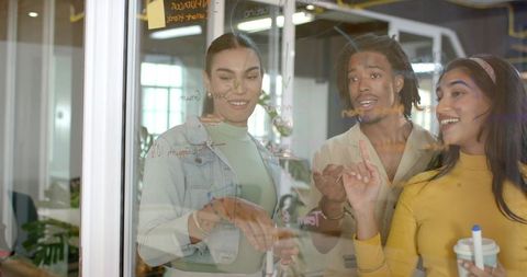 Multicultural team brainstorming at glass wall, planning ideas in coworking workspace