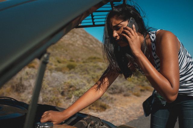 Woman talking on phone while checking car engine during summer road trip on country highway by coast. Useful for travel blogs, car maintenance tips, emergency roadside assistance services, and summer vacation planning.
