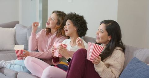 Three friends laughing and eating popcorn on cozy sofa during movie night at home